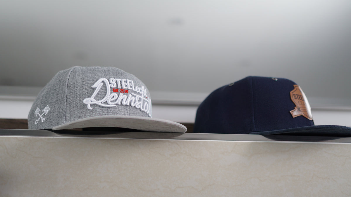 Zwei Snapback-Caps auf einer Ablage im Bad, die linke ist grau und die rechte dunkelblau